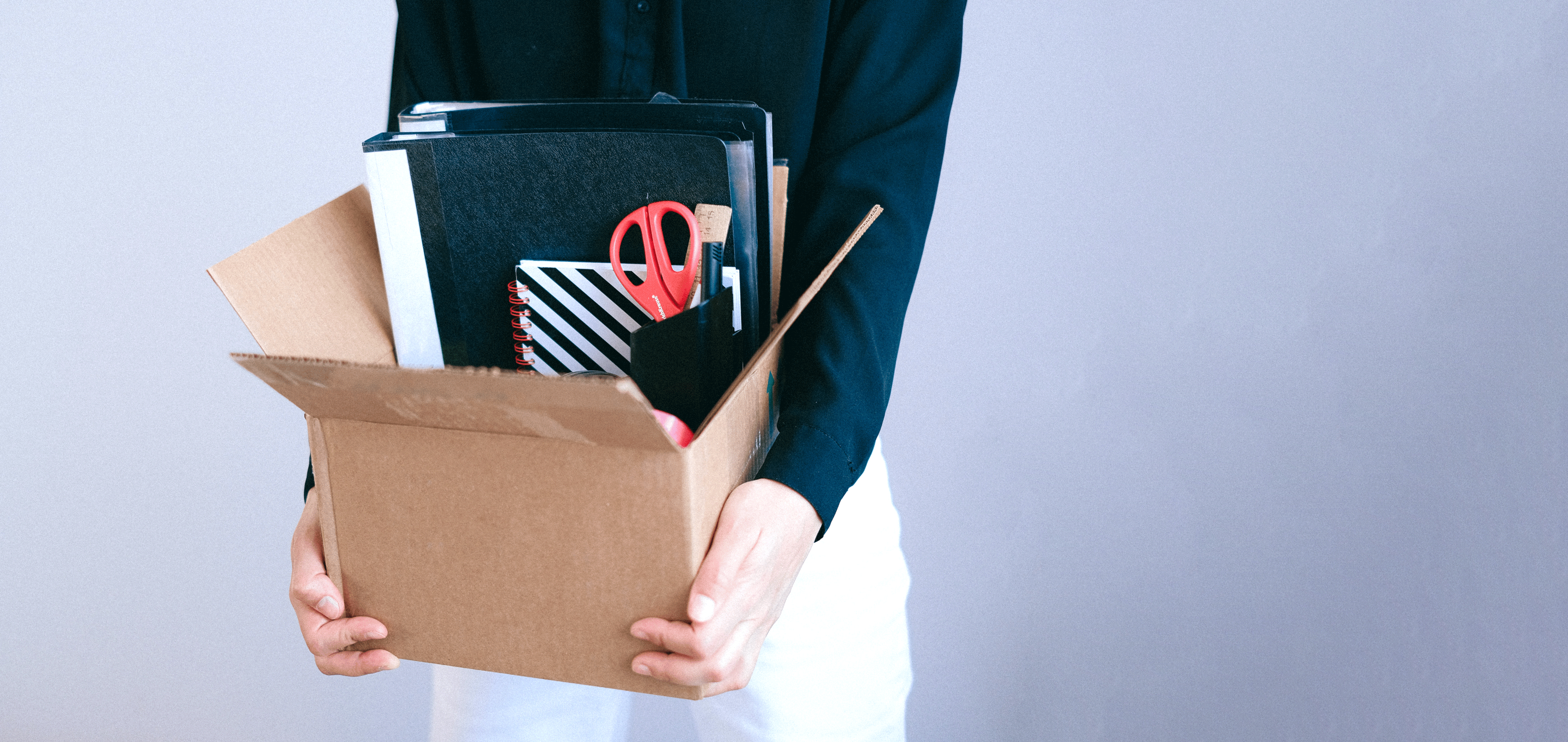 Person holding a cardboard box with red scissors, papers, pens, and various office supplies